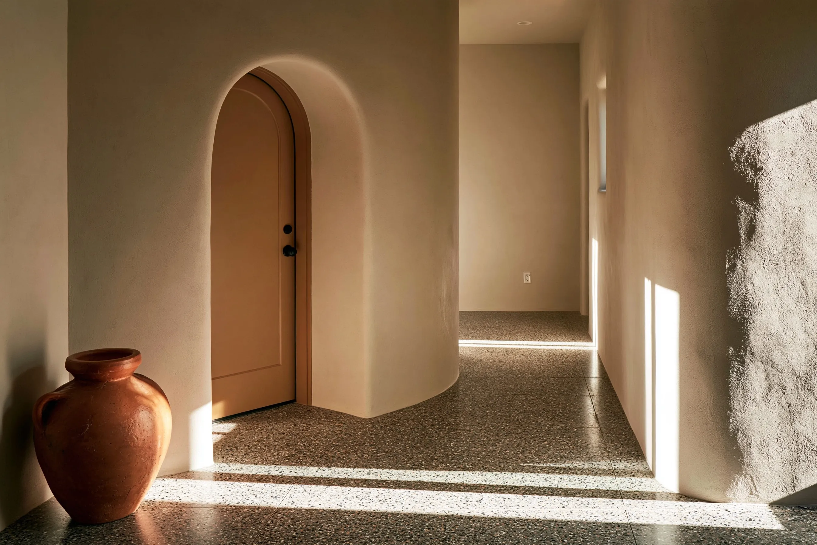 Entry hall and corridor with a salt-and-pepper fine-aggregate polished floor, an arched stucco doorway and a terracotta pot in daylight